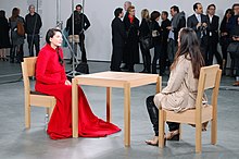Dwie kobiety siedziały twarzą w twarz przy stole, jedna z nich w dużej czerwonej sukience.  W alejce obok niej przechodzą ludzie.