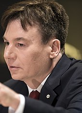 Ein Nahaufnahmefoto von Mike Myers, der einen schwarzen Blazer mit einer roten Krawatte trägt.Er scheint auf etwas zu zeigen.