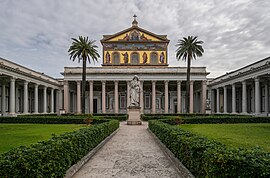 A courtyard with palm trees. And a greater-than-lifesized statue of Saint Paul holding sword in front of the colossal portico of the basilica and a large mural covering the upper facade