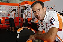 Smiling clean shaven middle aged man wearing a white shirt leaning over a motorcycle to right with other males wearing similar team shirts in background