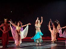 A blond female performer. She is wearing a Bollywood-inspired outfit, while surrounded by a group of dancers.
