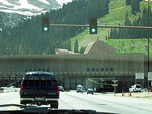 An automobile is about to pass under a traffic signal on a road leading to an opening in a building against a mountain. On the roof of the building, large ventilation hoods are visible.
