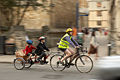 Дорослий на велосипеді і двоє дітей на двоколісному двомісному веловізку