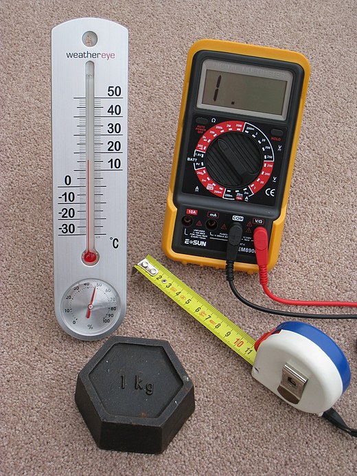 A kilogram mass and three metric measuring devices: a tape measure in centimetres, a thermometer in degrees Celsius, and a multimeter that measures potential in volts, current in amperes and resistance in ohms.