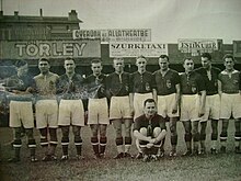Черно-белая фотография футбольной команды.  Десять игроков стоят, выстроились в ряд, одиннадцатый сидит.