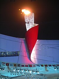 four pillars with flame at their tops surrounding a single fifth pillar in the middle, also with flame at the top. The background is sky with mountain.