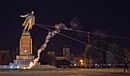 Die Lenin-Statue, an der zwei Stahlseile befestigt sind, wird in der Dunkelheit von dem Podest gezogen. Im Vordergrund eine kleine Rauchsäule, im Hintergrund eine große Menschenansammlung