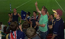 Gruppe von Fans, die für ihr Team in Stadionständen singen, die mit australischen Flaggen und verschiedenen Fußballutensilien geschmückt sind.