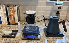 A set-up used to brew coffee, featuring (from left to right) a coffee dosing tray on a small scale, a small spritzing bottle, a V60 pour over with paper filter on a digital scale, a gooseneck kettle, and a coffee grinder. Coffee Pour Over Setup.jpg