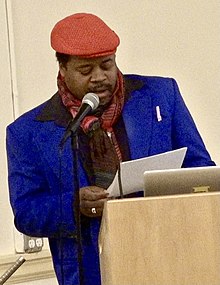 Tate at a podium, reading into a microphone; he wears a blue coat and orange hat