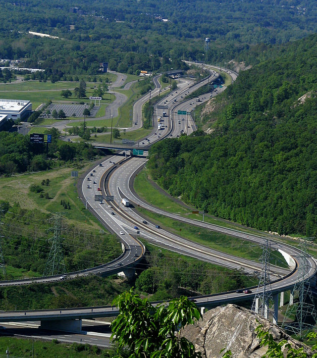 A view of a freeway leaving an interchange from a mountaintop, heading between development to the left and mountains to the right