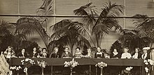 Ein Gruppe von dreizehn Frauen auf einem Podium sitzend