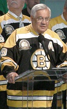 Photo de Bucyk portant le maillot numéro 9 des Bruins et qui s'adresse à un public depuis un pupitre avec un micro.