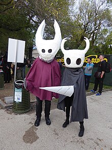 Photographie de deux personnes déguisées en personnages du jeu, avec masques, capes, et arme factice.