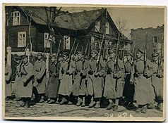 Photographie en noir et blancs de soldats de l'Armée rouge défilant alignés, tenant chacun un fusil, avec derrière eux un bâtiment en bois.