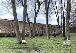 Im Vordergrund ist ein Weg und ein Wiesenabschnitt zu sehen. Mittig vom Bild befinden sich zwei Bäume, an deren Füßen die beige Gedenkkugel liegt. Den Hintergrund füllt die braune Lagermauer aus, an der verschiedene Gedenktafeln angebracht sind.