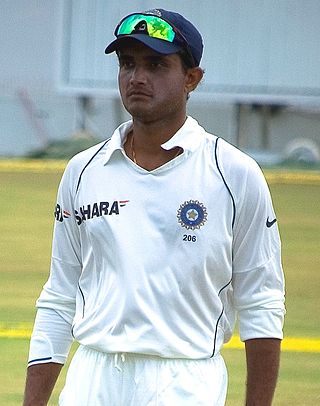 A middle-aged man stands to wear a white long-sleeved shirt and white trousers, while he has sunglasses resting on a cap that is on his head. Green grass and a boundary line are in the background.