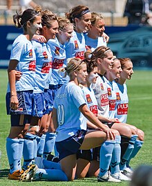 Um grupo de mulheres, vestindo conjuntos de futebol azul celeste, em duas fileiras.