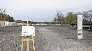 Der Wegweiser aus Holz trägt ein weißes Schild mit Hinweisen zur Ausstellung. Im Hintergrund ist das Gelände der Mahn- und Gedenkstätte Ravensbrück zu sehen, das mit einem steinartigen, grauen Material bedeckt ist. Einzelne Gebäude sind erkennbar.