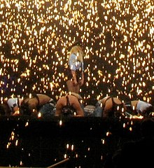 Distant image of a blond woman. She is standing on an elevated stage, wearing a policewoman uniform and carrying a black baton at the height of her legs. Several people are around her in kneeling position, the males shirtless and wearing jeans and the females wearing similar policewomen uniforms. A rain of sparks is falling over their heads.