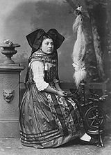 Fotografi fra 1870-tallet av kvinne fra Alsace kledt i folkedrakt ved en liten rokk med høyt rokkehode med mye ullfiber.