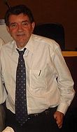 A man standing with his hands in his pockets. He is wearing a white shirt with a dark patterned tie.