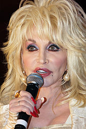 A late-middle-aged Caucasian woman with voluminous blonde hair and heavy make-up is singing into a microphone.