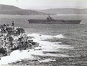 Grainy photograph of an aircraft carrier manoeuveing around a cliff-faced headland with a lighthouse on top. More landmasses are visible in the background.