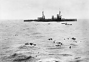 A large warship lowers boats to pick up sailors floating in the open water
