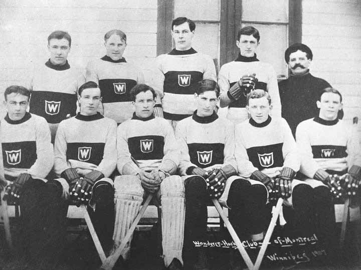 Black and white image of eleven men posing for a photo， with six of them sitting on a bench， and the other five standing behind them. All but one man are wearing the same wool sweater， white with a dark band around the torso; within the band is a small white 