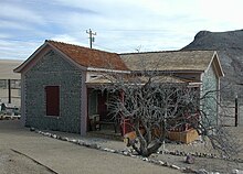 A one-story house with a relatively new roof sits on a manicured bed of gravel. The house and its tidy porch are at least partly made of wood, but the outer walls appear to consist of hundreds of round objects embedded in a masonry matrix. A short, leafless, gnarled tree grows in front of the house along a tidy gravel sidewalk. A barren hill rises in the middle distance.