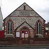 Vorderansicht einer winzigen, schlichten Kapelle aus unregelmäßigem Feuerstein und roten Ziegeln.  Eine dunkle Steintafel unter einem Schlitzfenster nahe der Dachlinie liest "WESLEYAN CHAPEL".  Ein Eingangsportal wird von zwei breiten, niedrigen Fenstern mit Spitzbögen flankiert.