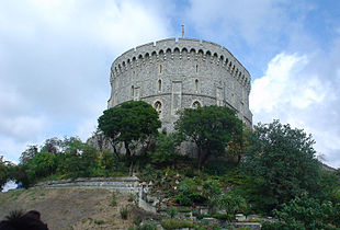A photograph of a white and grey stone tower, on a tall mound of earth dotted with small trees and bushes.