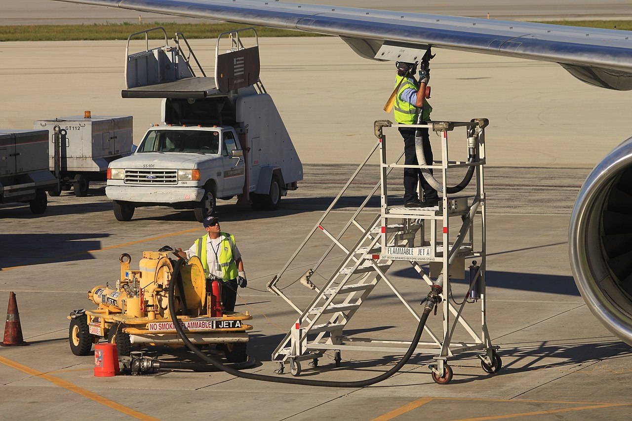 Airport ground staff are refueling an aircraft while standing on a ladder on a sunny day.