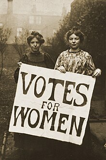 Two woman carry a sign reading „Votes for Women“.
