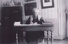 Photographie représentant une femme âgée écrivant à un bureau.