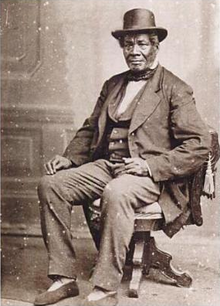 A black-and-white photograph of an African-American man in a hat and suit seated in a decorative wooden chair.
