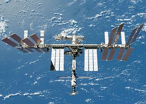 A planform view of the ISS backdropped by the limb of the Earth. In view are the station's four large, gold-coloured solar array wings, two on either side of the station, mounted to a central truss structure. Further along the truss are six large, white radiators, three next to each pair of arrays. In between the solar arrays and radiators is a cluster of pressurised modules arranged in an elongated T shape, also attached to the truss. A set of blue solar arrays are mounted to the module at the aft end of the cluster.