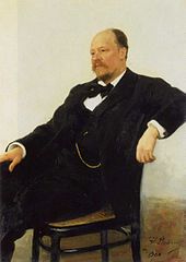 A balding man with a mustache, wearing a dark suit with a bow tie and sitting in a chair.