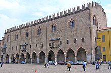 Un lungo edificio in pietra grigia con una fila di archi a livello del suolo, sopra il quale si trova una fila di piccole finestre intervallate da balconi.  Sopra questa fila c'è una linea di finestre più grandi sotto una linea del tetto merlato.