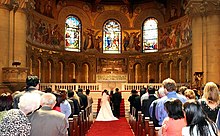 Looking down the center aisle at a bride and groom standing before a minister in front of the altar: a red carpet covers the floor of the aisle, and the church is full of on-lookers.
