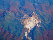 Fotografie color afișând, în inima unui lanț muntos împădurit, un crater vulcan văzut din cer.