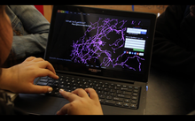 This is a picture of an open laptop on a desk. The student using the laptop is not pictured but you can see one hand on the keyboard and one hand on the mouse pad as if they are in the middle of using the computer. The website on the laptop says EyeWire in rainbow colors at the upper left of the screen and there is a menu option bar across the top of the webpage. The webpage has a black background with a large picture of what appears to be a neuron structure (which looks like branches of purple squiggly lines coming from a small, spherical component). To the right of the screen is where you enter login information and the top left it says, "What is EyeWire? Play a game to map the brain."