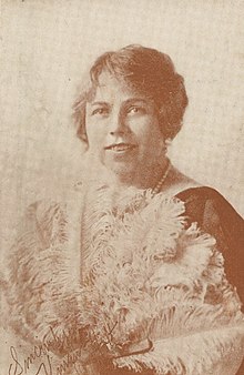 A sepia-toned portrait of Vivian Holt, a smiling white woman with light eyes and wavy hair, holding a feather fan