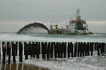 A ship sailing just in front of the beach. From the ship a dark jet of sand and water is blown towards the coast
