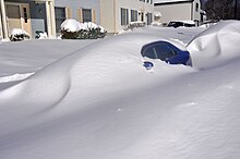 Only in the area of ​​the rear door and the fender is visible blue car under a long, man-high snowdrift.  Houses can be seen in the background.