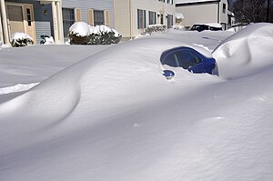 Ein grüner Lastwagen, der einen gelben Schneepflug inmitten fallenden Schnees schiebt.