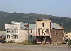 In the background, a large forested hill, increasing in height to the left, fronts a blue-gray sky. In the foreground, a road with no markings is at the base of the photograph, on the other side of which are four buildings. The leftmost building is obscured by a fence, and both obscured by the building to its right. That two-storey building has wooden steps with a white railing leading to the front door, on either side of which are windows. Above these is another row of windows, and above those the name "The Front St. Inn". The next building has the signage "Yukon Hotel" above two windows flaking a balcony door. The cream-coloured facade of the building matches the balcony railing, which is secured by four burgundy posts. Below the balcony is the front entrance, with cream-coloured doors flanked by large windows. The right side of the log building is visible extending to the background to the right, four equally spaced windows on each floor. The fourth building is partially behind and to the right of the Yukon Hotel.