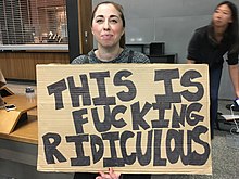 A woman holding a cardboard sign that reads "THIS IS FUCKING RIDICULOUS"