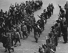 Several lines of school children march diagonally from top right to bottom left. Each carries a bag or bundle and each raises their right arm in the air in a salute. Adults stand in a line across the bottom right hand corner making the same gesture.